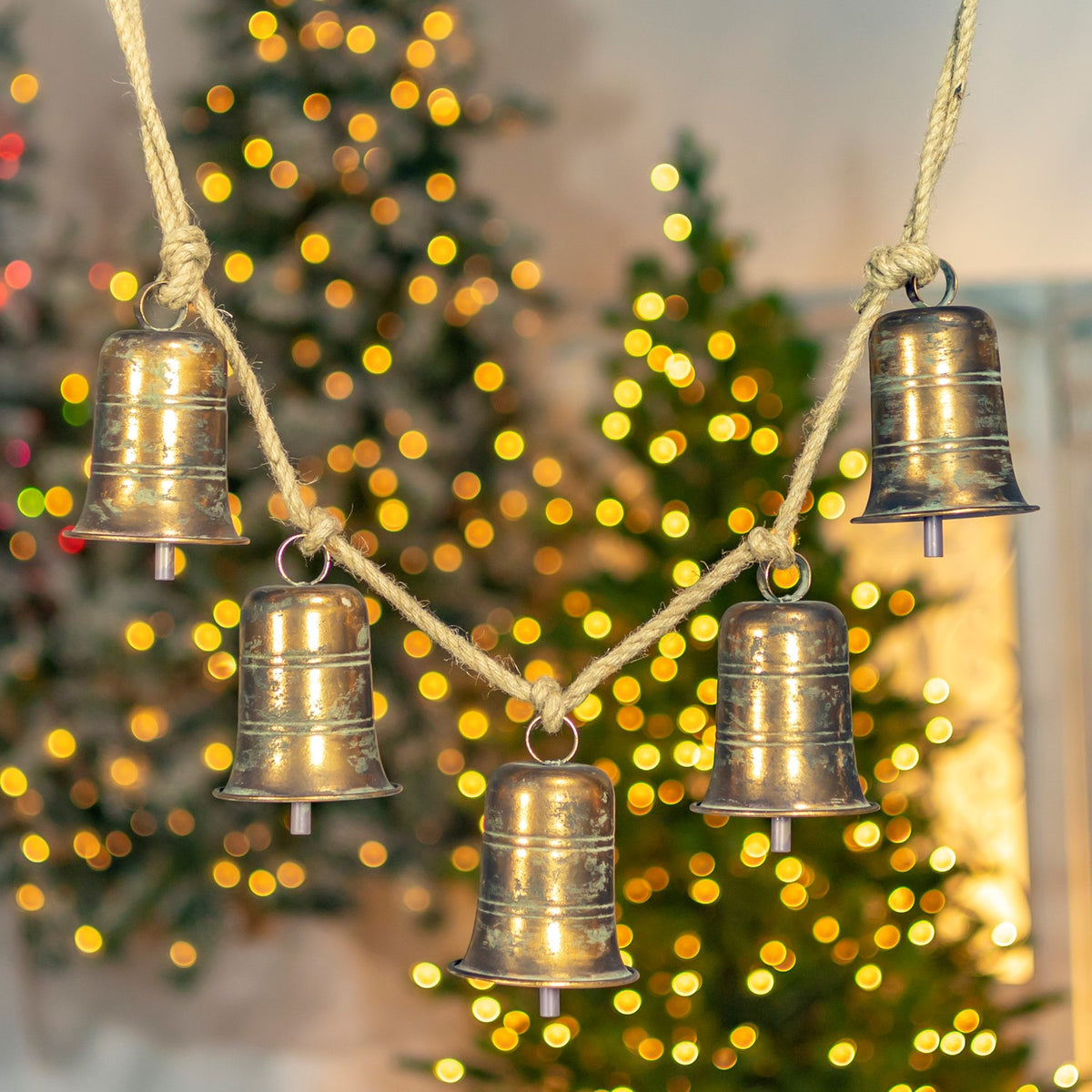 Five antique-gold bells with horizontal bands hang from a rope against a glowing Christmas tree.