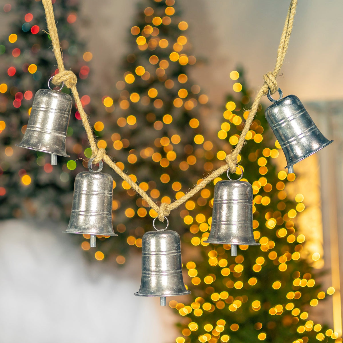 Five smooth, flared silver bells hang from a twine strand in front of blurred holiday lights.