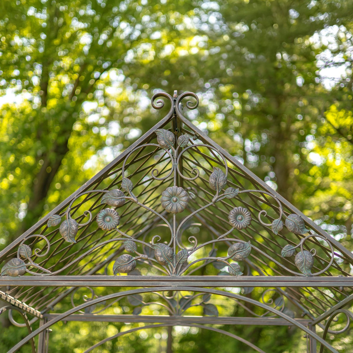Peak of an iron garden arch with curled scrollwork, leaf shapes and flower ornaments against trees.