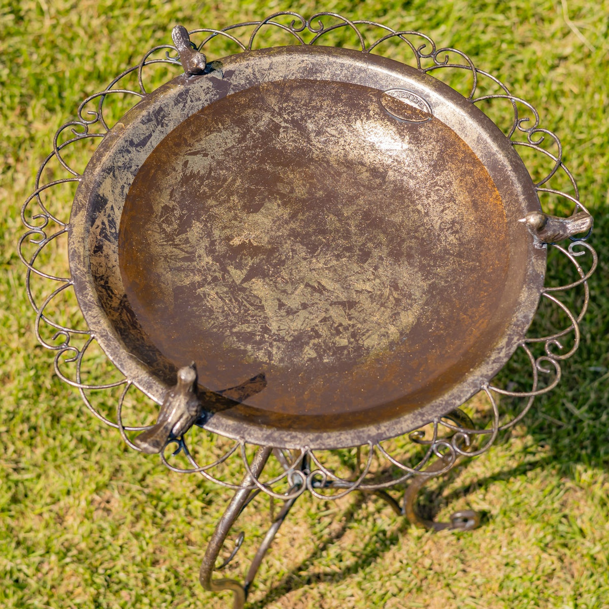 Overhead view of a round, weathered metal birdbath with three small bird figures and an ornate scrollwork rim.