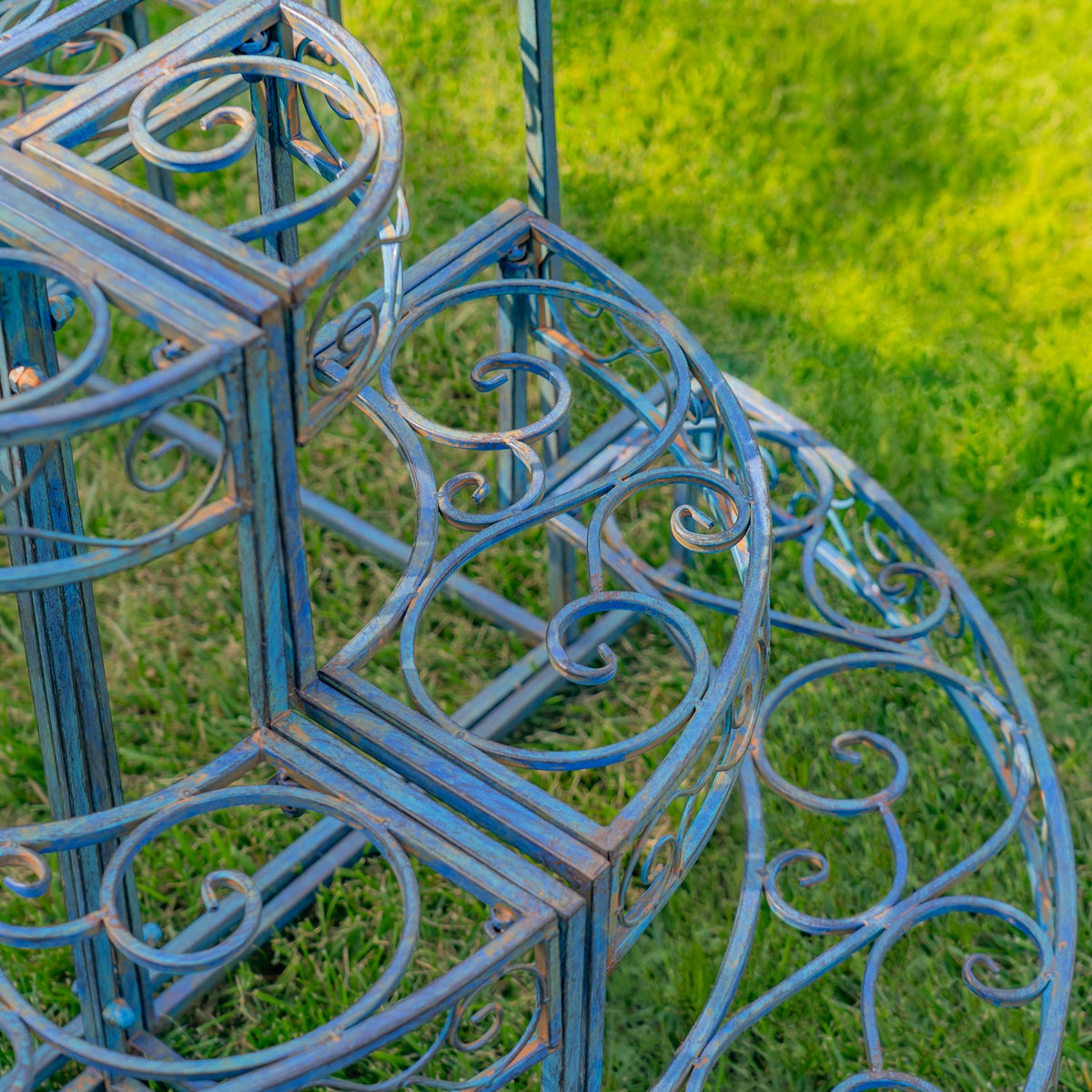 Close-up of the plant stand’s blue metal scrollwork and curved end rail above the grass.