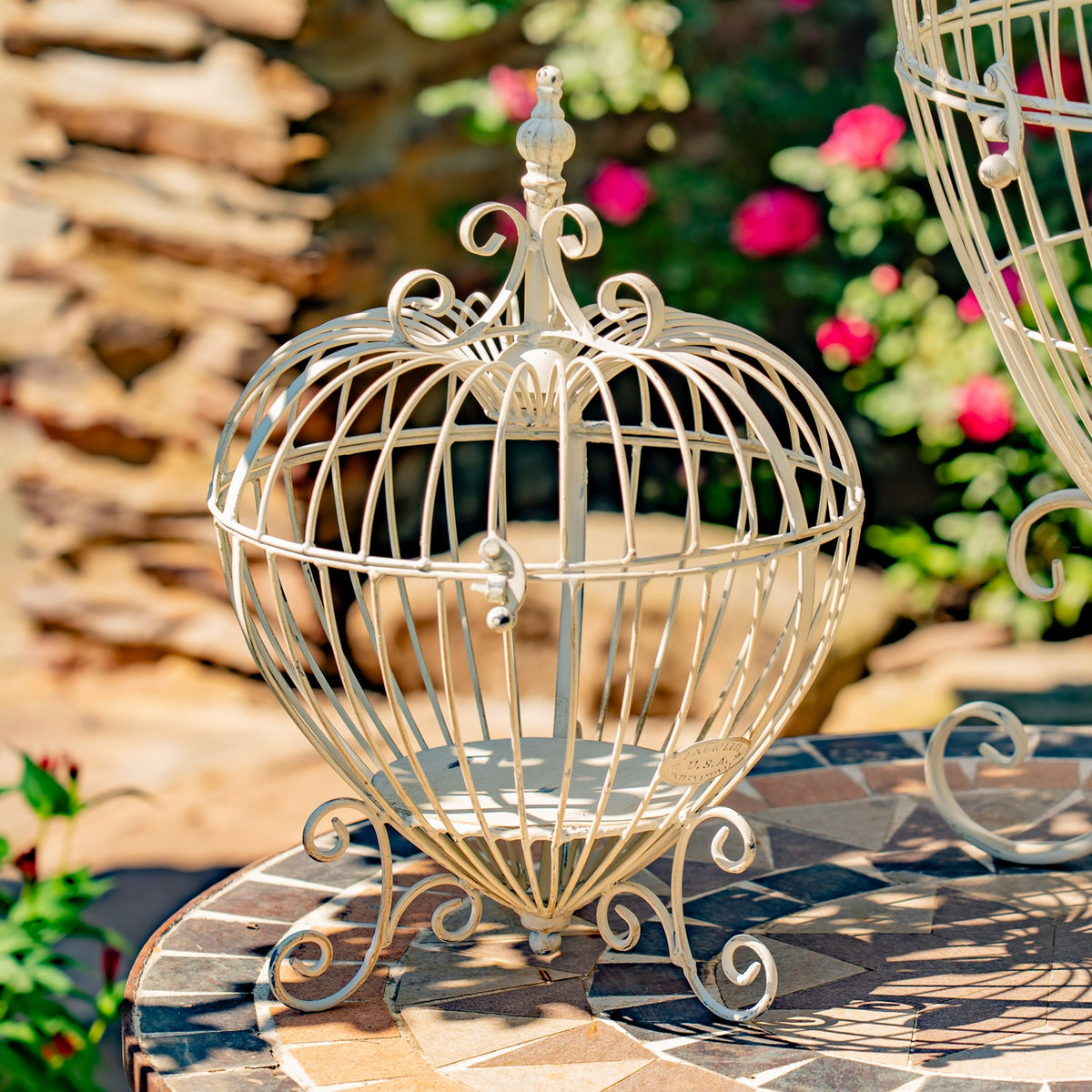 Ornate heart-shaped wire plant stand on a tiled table in a flower garden.