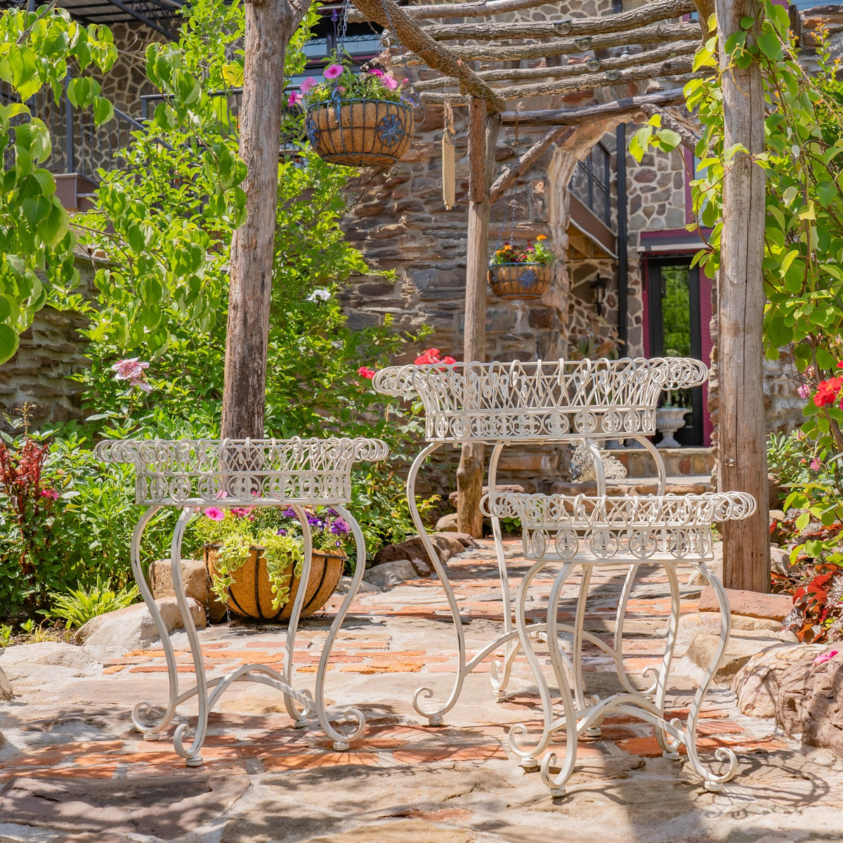 Three pale metal plant stands arranged on a stone patio, with flowers and greenery around them.