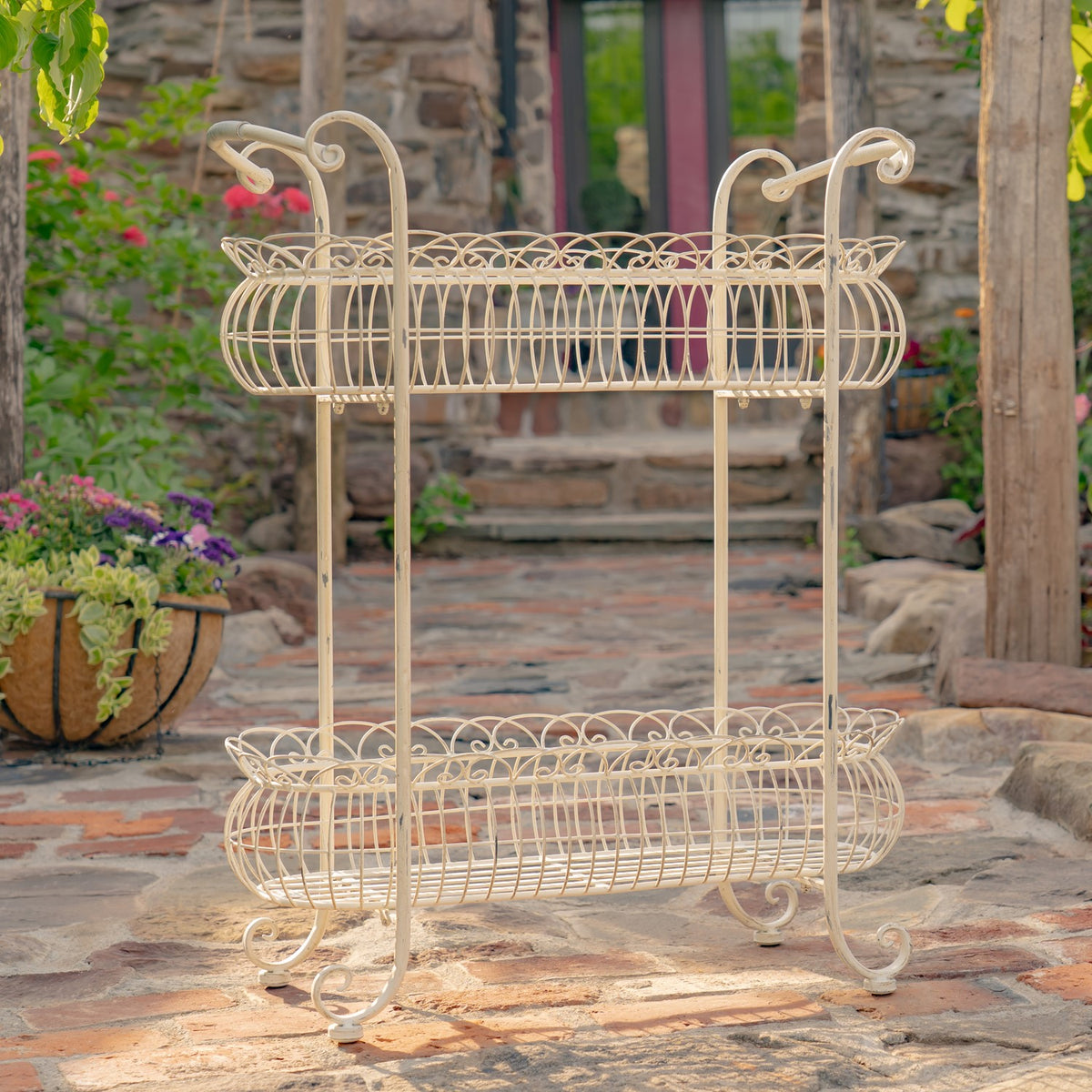 Full view of the two-tier wire plant stand on a stone patio, with a stone wall and potted plants behind it.