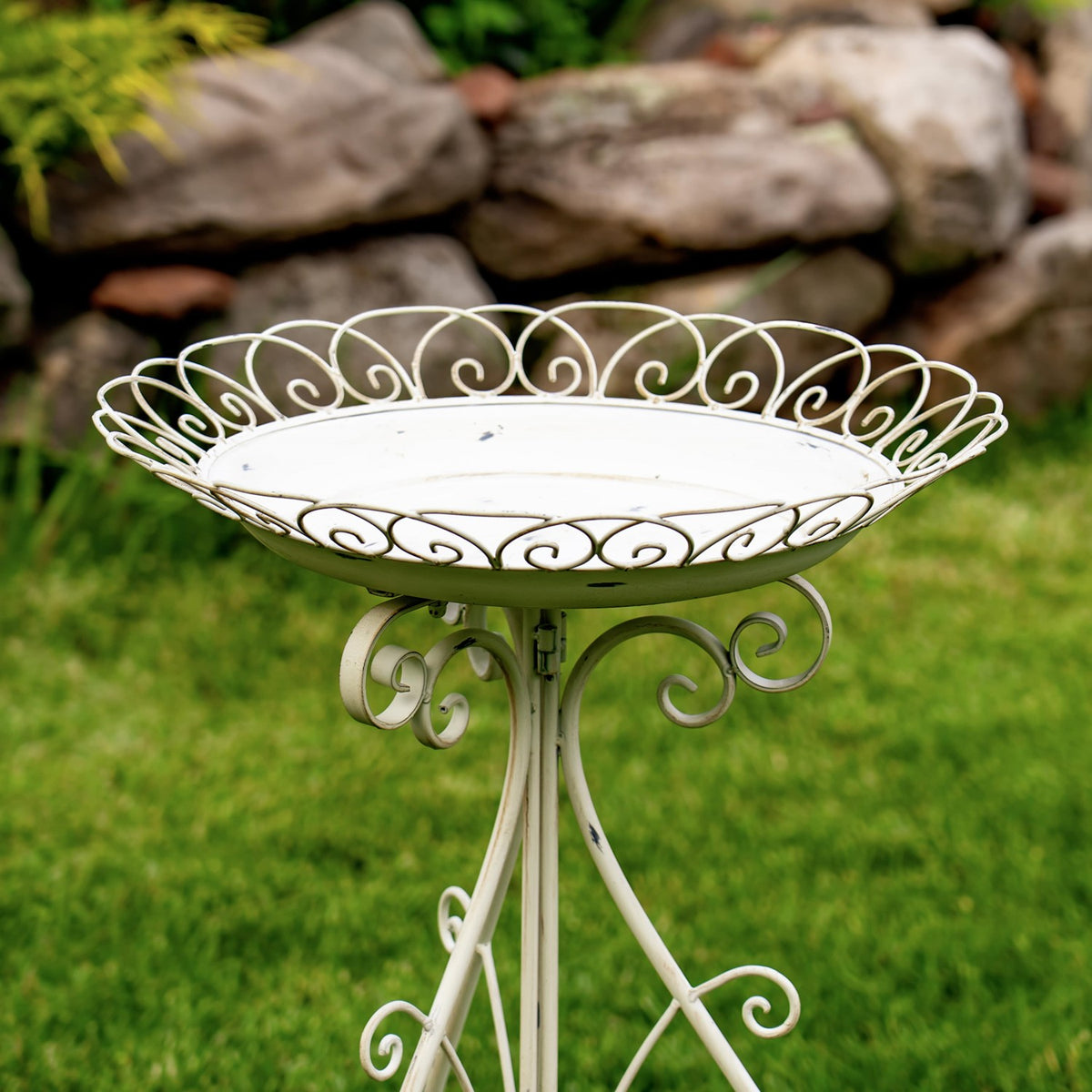 White round birdbath with ornate scrollwork rim and folding iron stand on a lawn.