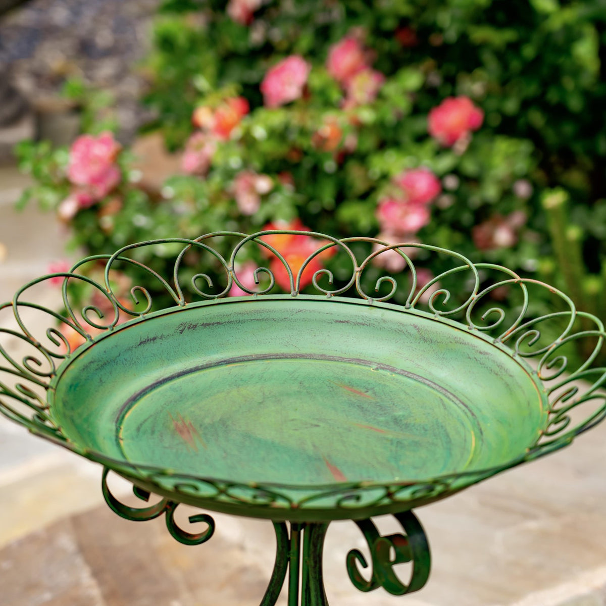 Green round birdbath with an ornate scrollwork rim and folding iron stand, set among flowers.