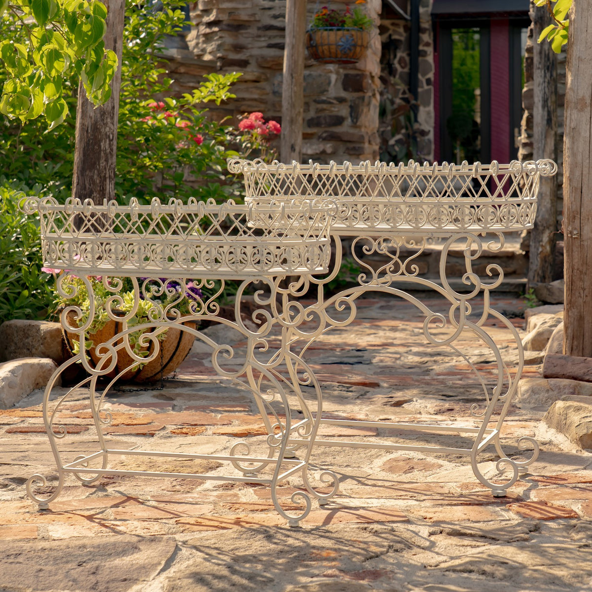 Two ornate ivory metal plant stands with oval baskets arranged on a stone patio.