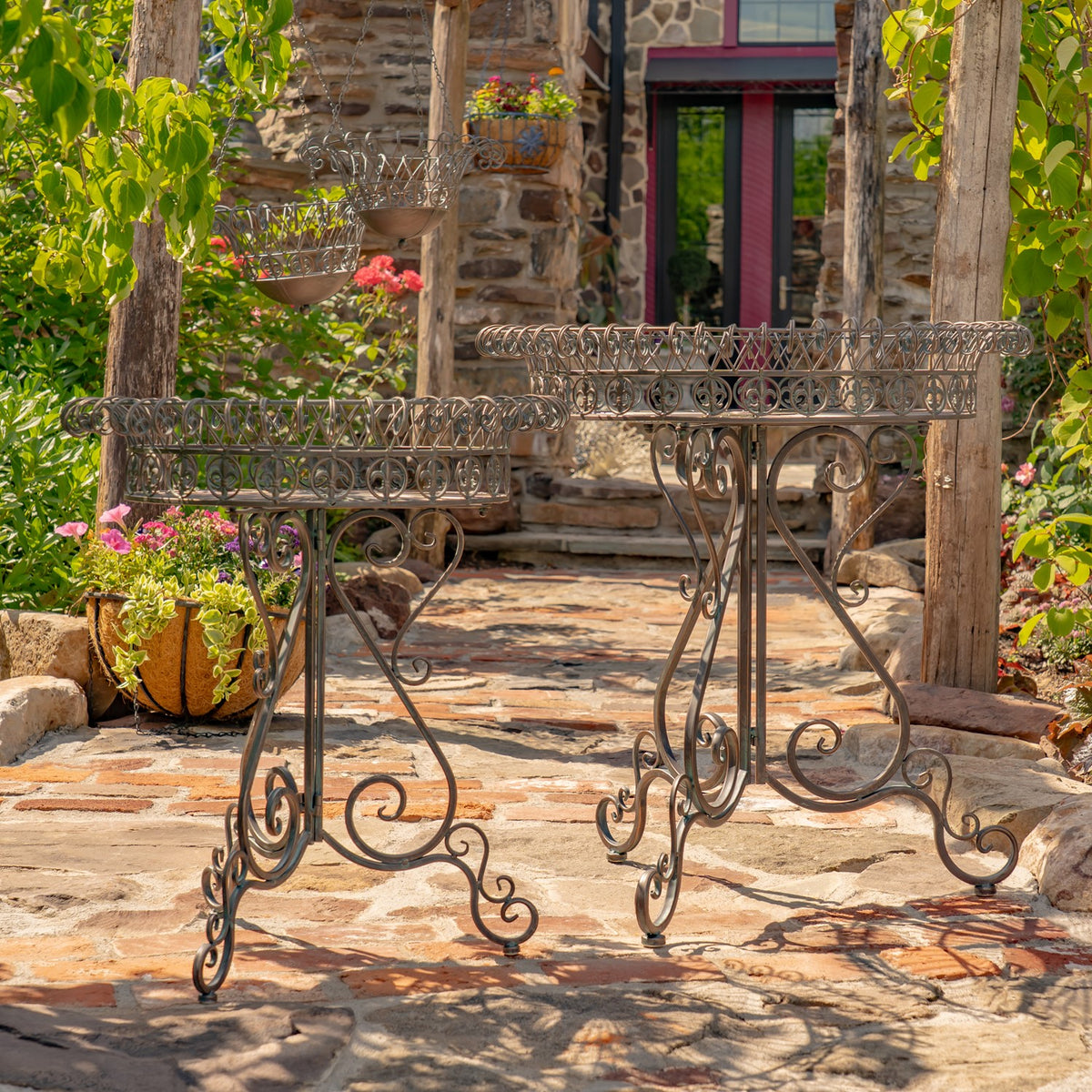 Two ornate round iron plant stands with wire basket rims sit on a stone patio.