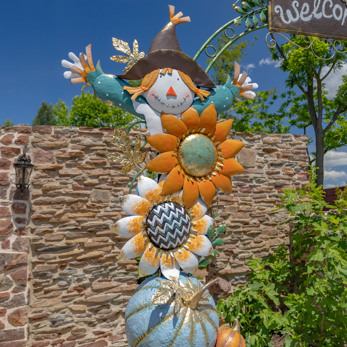 A scarecrow figure and layered sunflower and daisy decorations rise above stacked blue and orange pumpkins.
