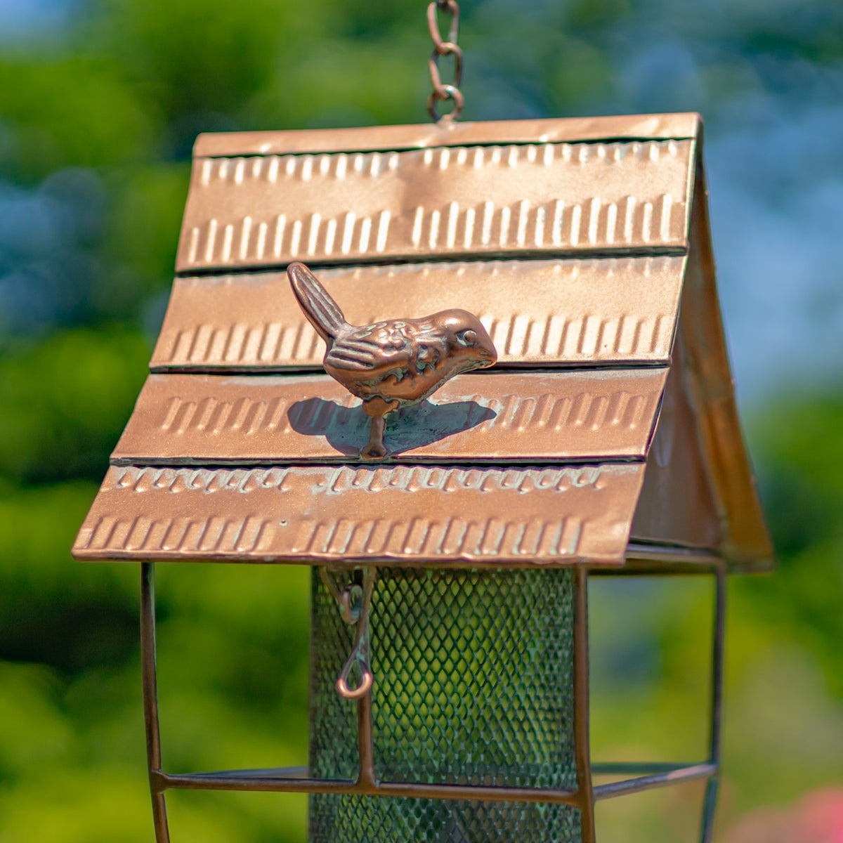 A small bird figurine sits on the ridged metal roof above the feeder’s mesh hopper.
