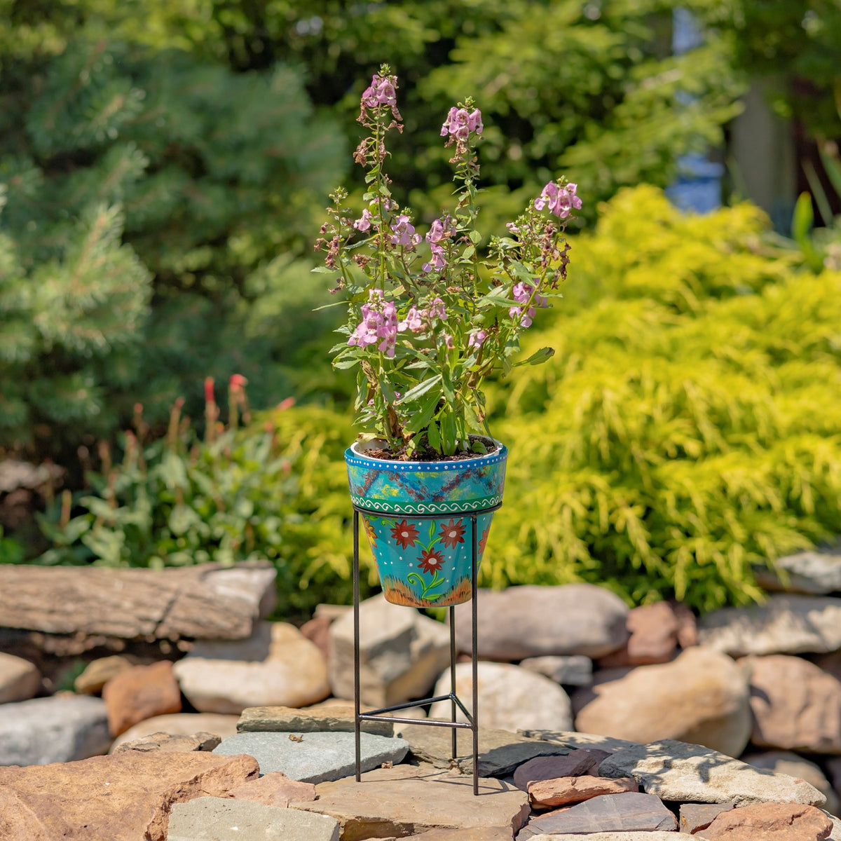 Turquoise flowerpot with bright painted floral patterns on a slender iron stand in a garden.