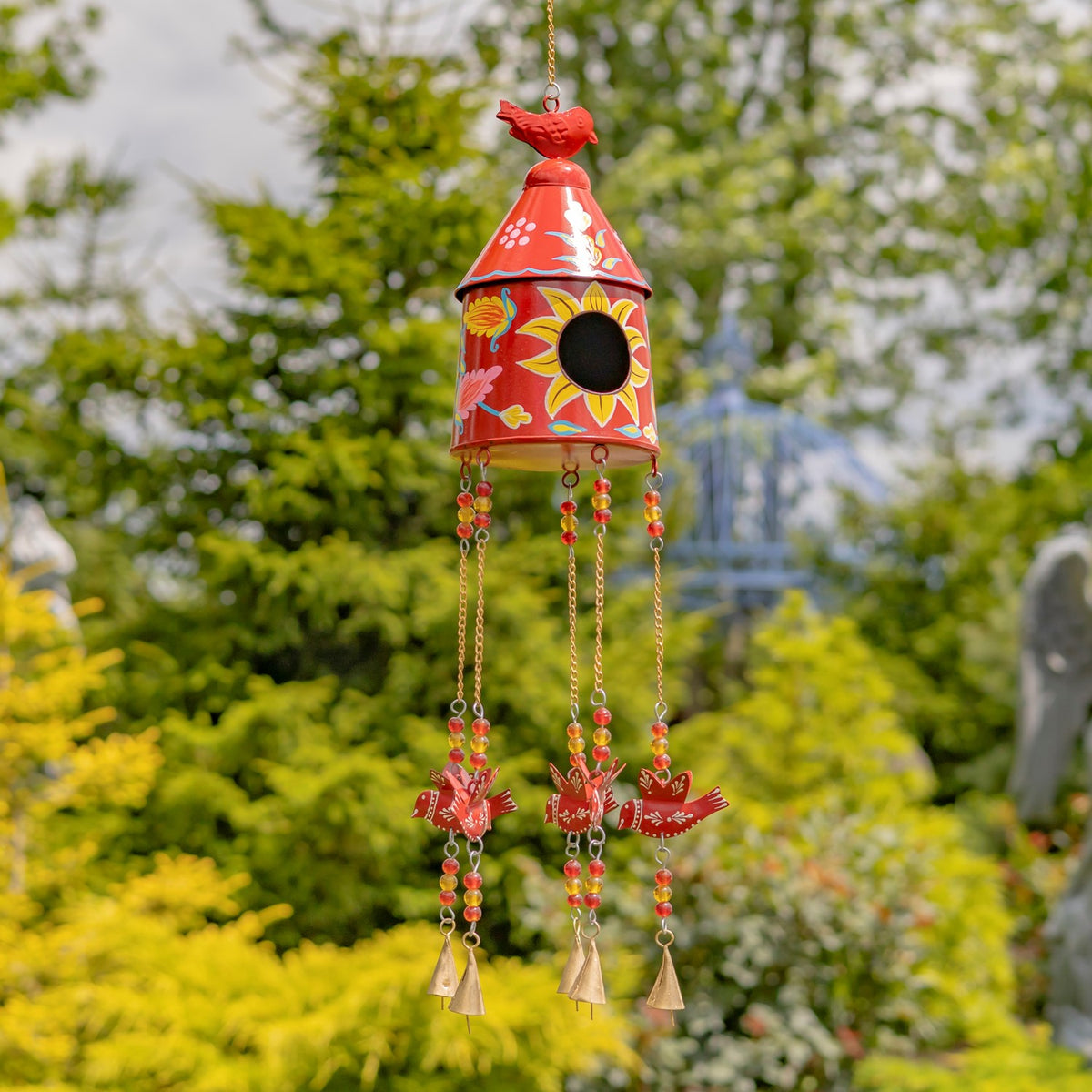Red birdhouse wind chime with painted floral details, colorful beads and dangling bells in a garden.