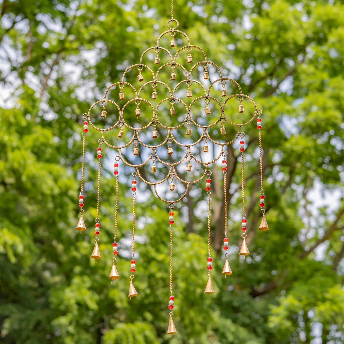 Gold ring-shaped wind chime with red and white bead accents among green trees.
