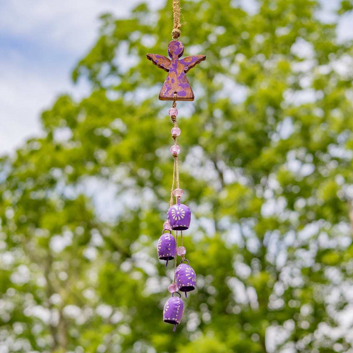 A purple angel wind chime with pale beads and purple dangling bells hangs in front of trees.