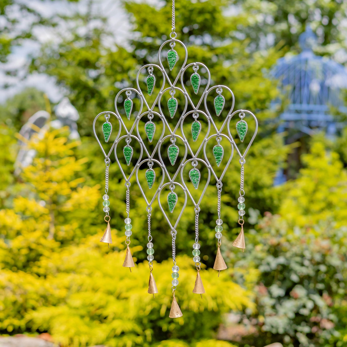 Silver-tone wind chime with green accents and dangling bell-shaped pieces hanging in front of garden foliage.