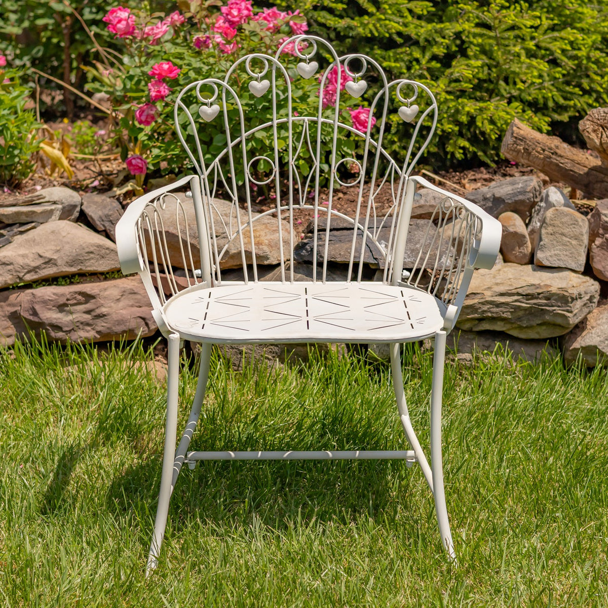 Pale metal garden armchair with curled scrollwork and heart accents on a lawn among flowers and rocks.