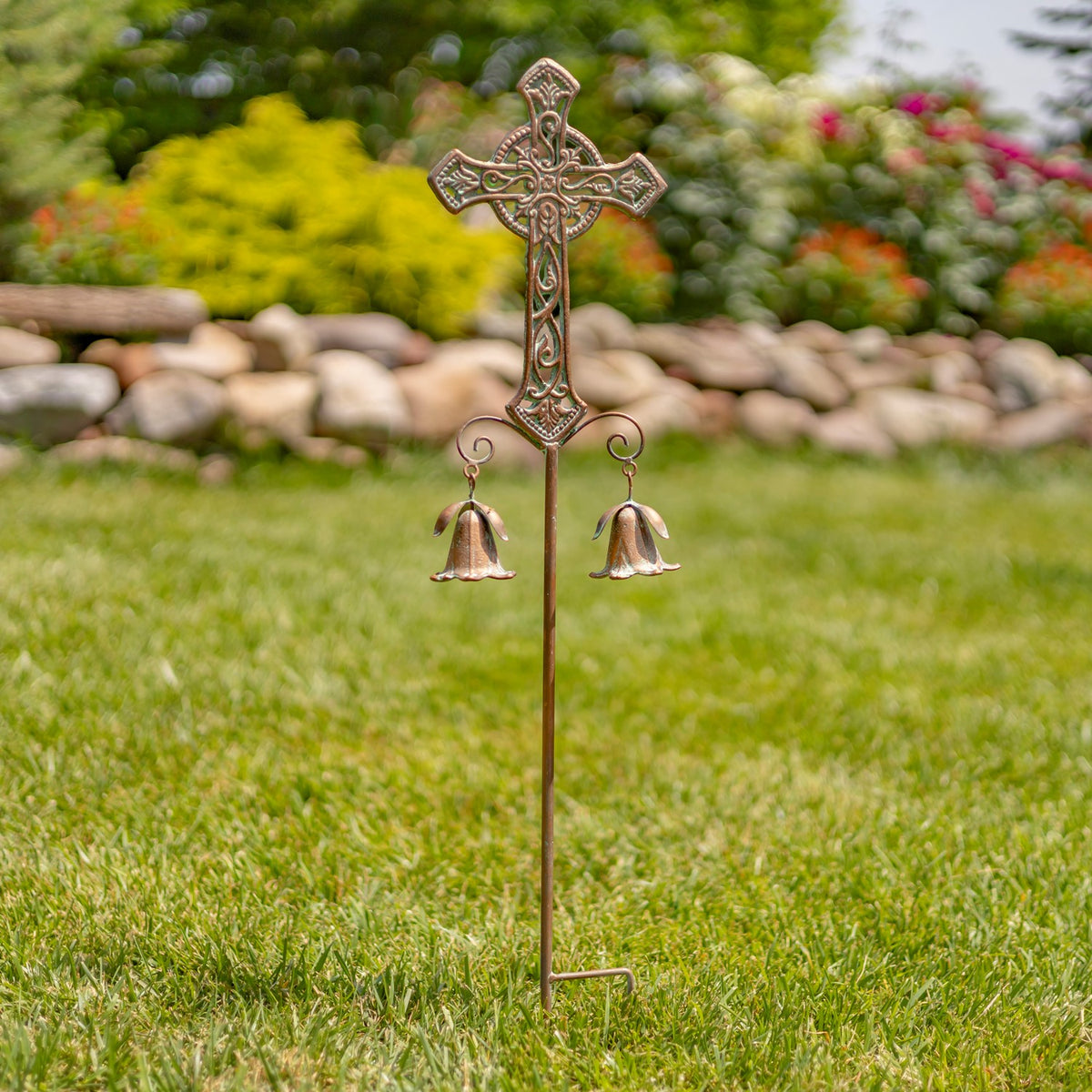 A dark, narrow cross design with hanging bells is shown planted in grass beside a stone border.