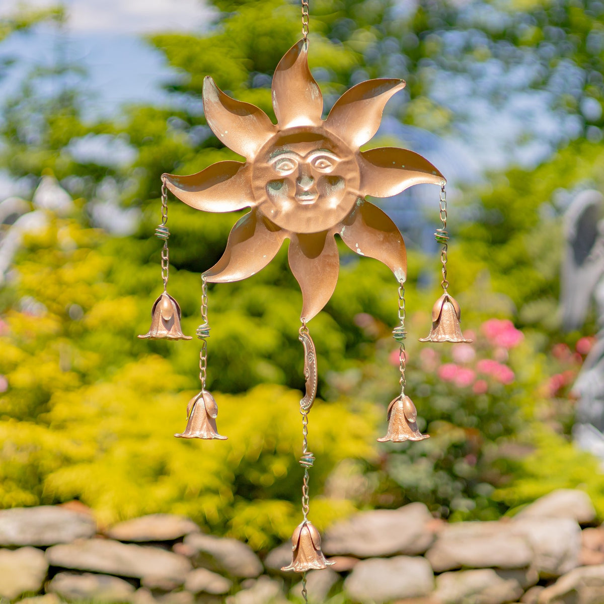 A sun-faced chime hangs against a garden backdrop, its rays surrounding the face and bells hanging below.