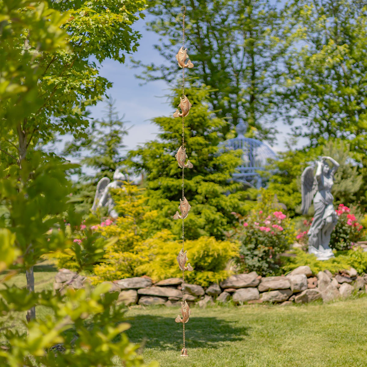 The fish-decorated rain chain hangs vertically in a garden between trees and stone edging.