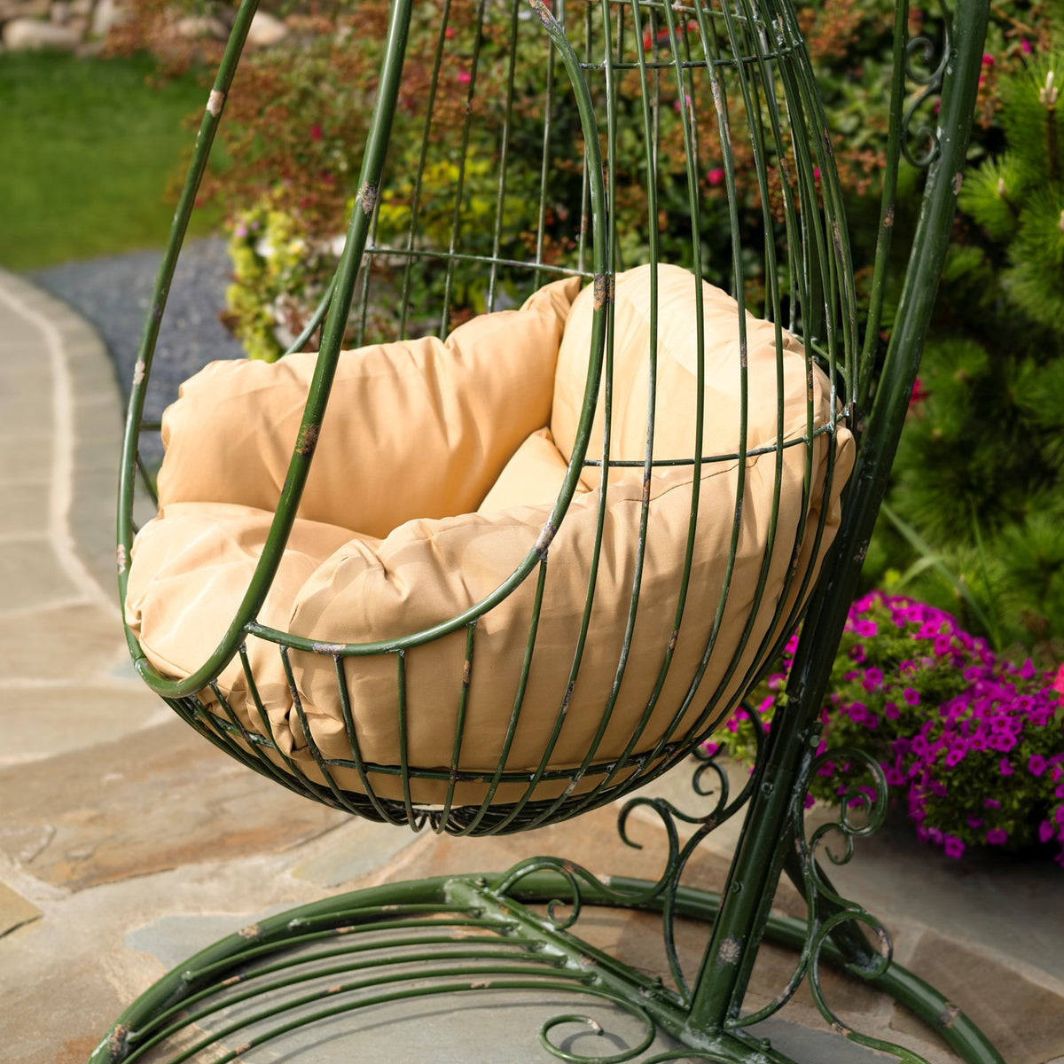 Green wire egg chair with a beige cushion, shown above its curved metal stand on a garden patio.
