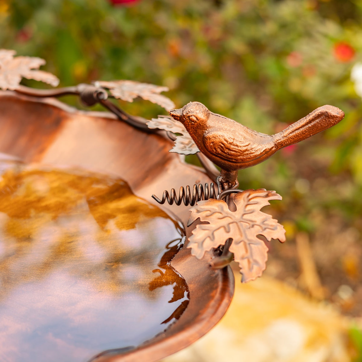 Close-up of a small bronze bird perched beside leaf-shaped accents on the rim of a birdbath.
