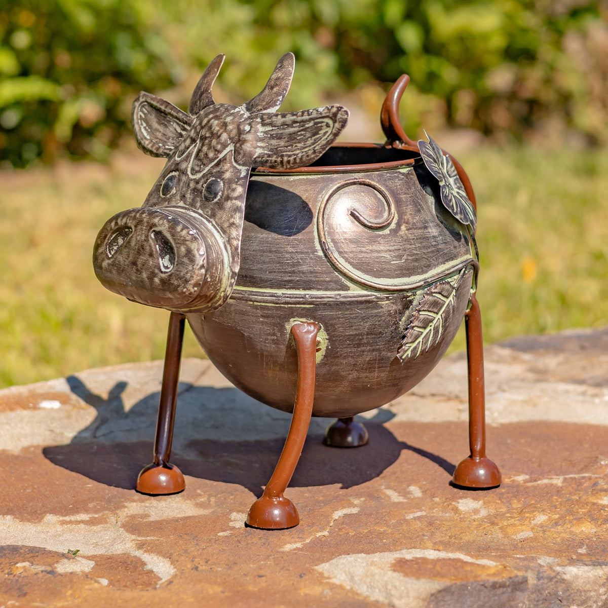 An empty cow-shaped metal planter with horns and a broad muzzle stands on long legs outdoors.