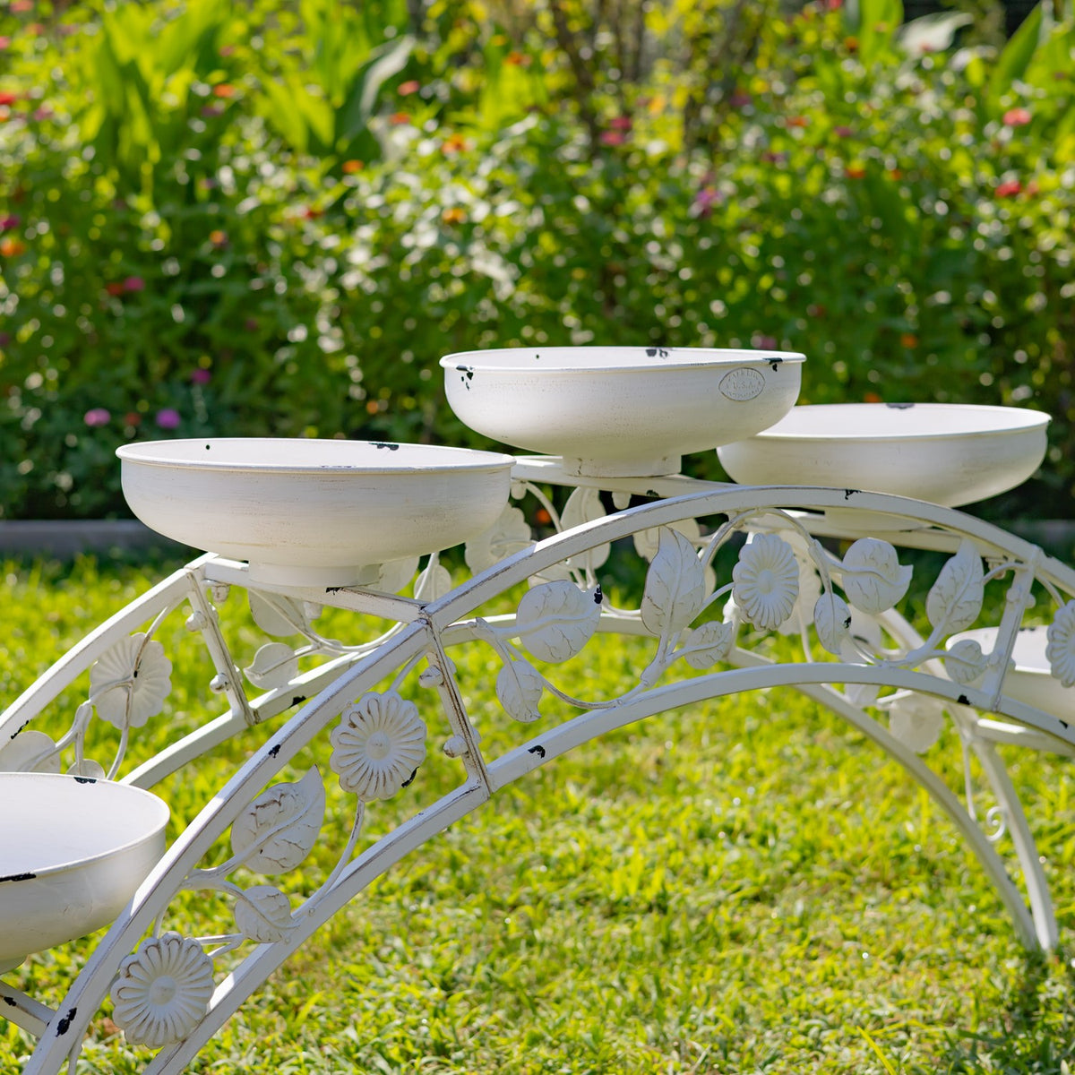 White arched metal plant stand with shallow bowls arranged at different heights on a lawn.