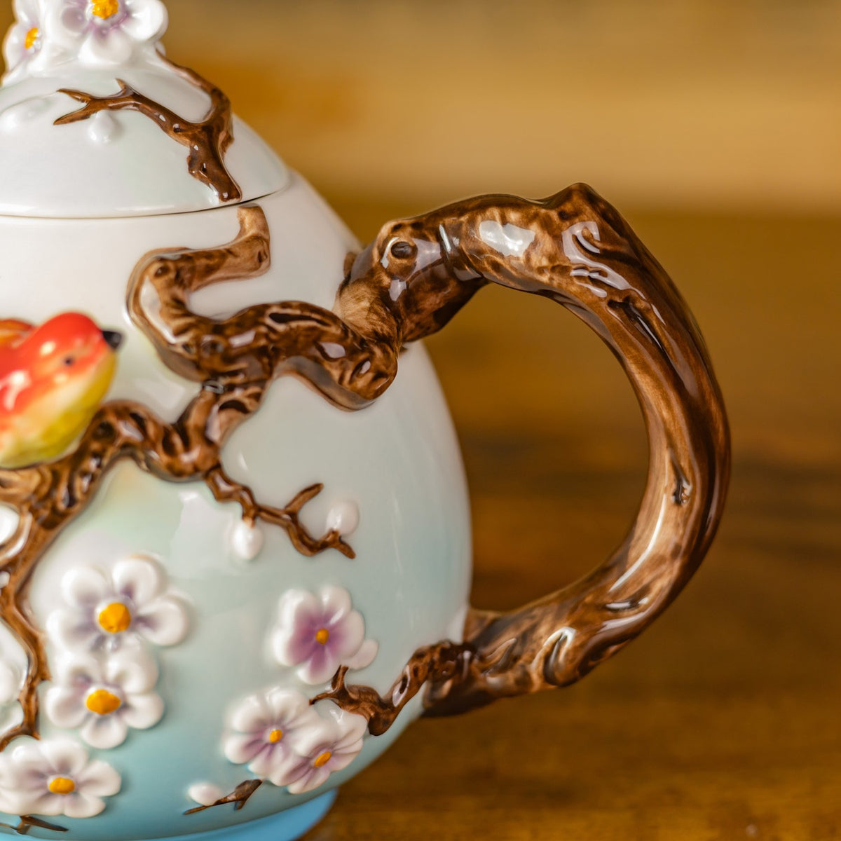 Sculpted brown branch handle curves beside raised blossoms on the teapot’s pale body.