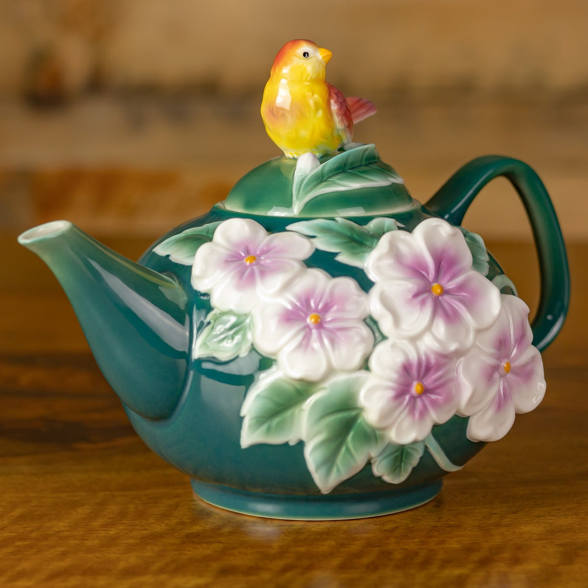Teapot shown from an angle, with its curved handle, pointed spout, raised flowers, and bird-topped lid.