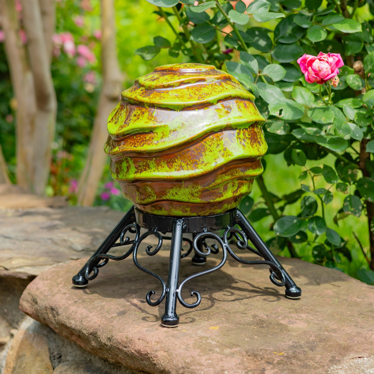 A ridged green and gold gazing globe on a black scrollwork stand, shown beside a blooming pink rose.