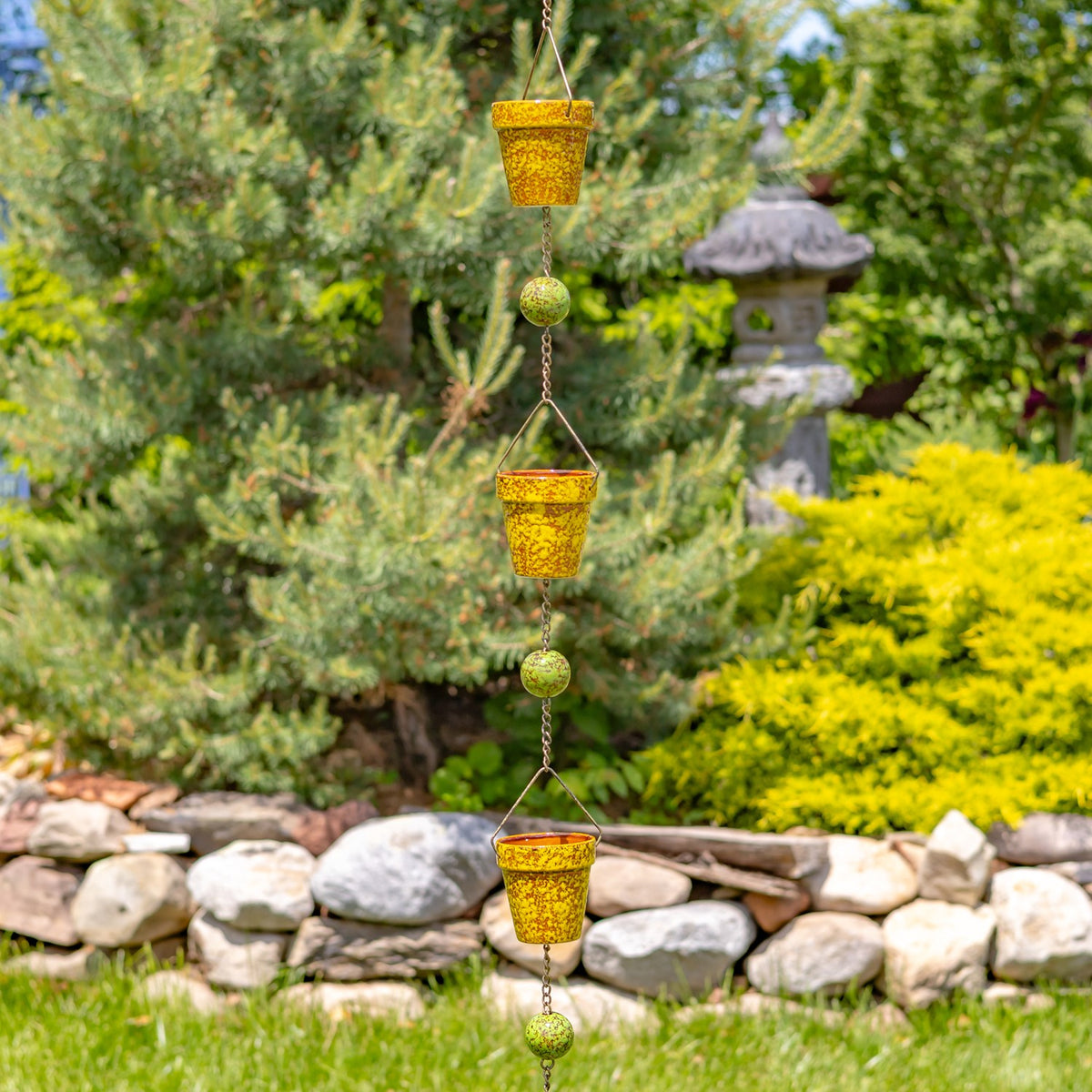 A vertical rain chain with three yellow speckled cups and green beads against a garden backdrop.