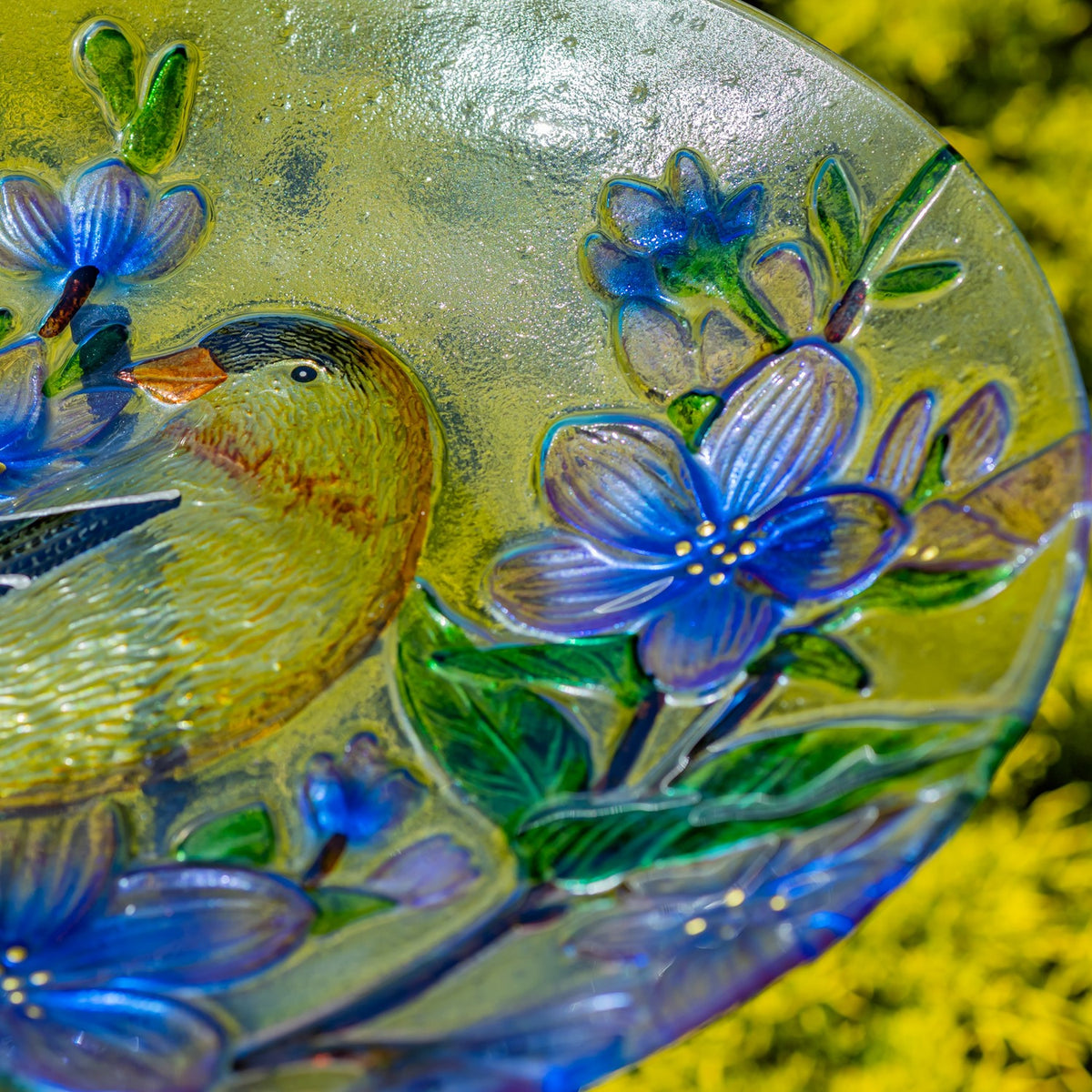 Close-up of a goldfinch beside a raised blue flower and green leaves on the glass bowl.