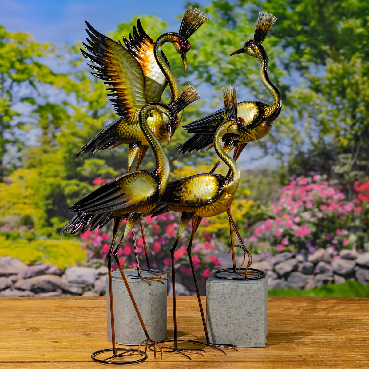 Metallic heron figurines stand on slender legs and circular bases beside planters on a garden table.