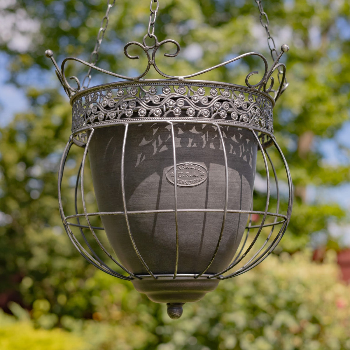 Ornate metal hanging planter with a dark inner pot, shown against a leafy garden.