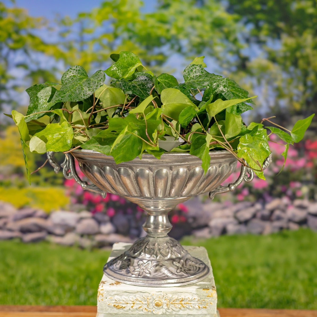 A silver urn filled with leafy greenery rests on a cream decorative pedestal.