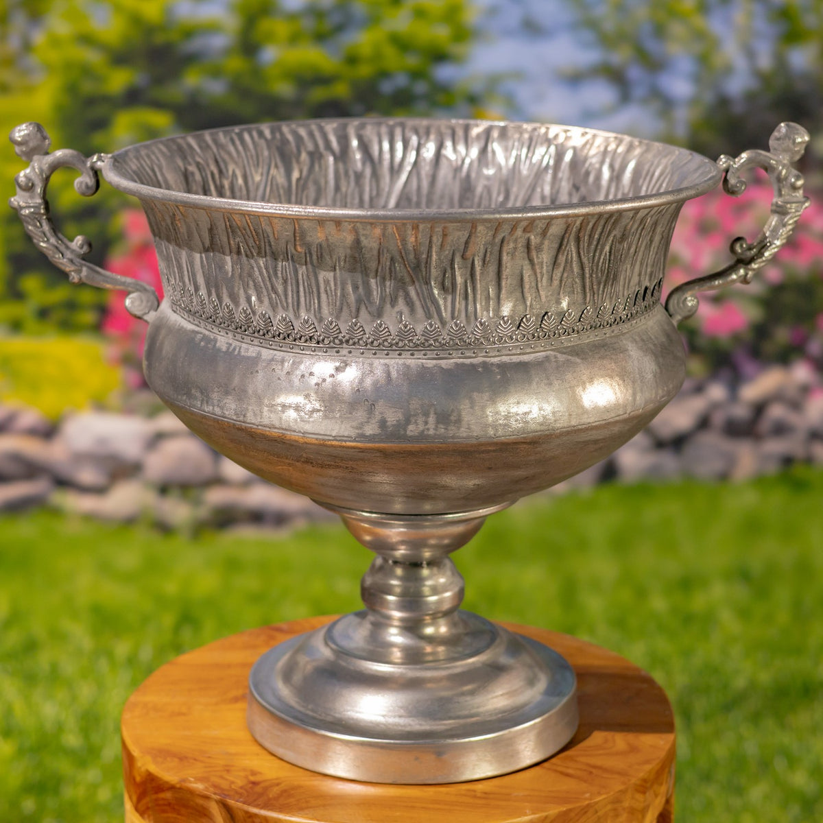 Close view of a silver urn showing its ribbed rim, embossed band, curved handles, and pedestal base.