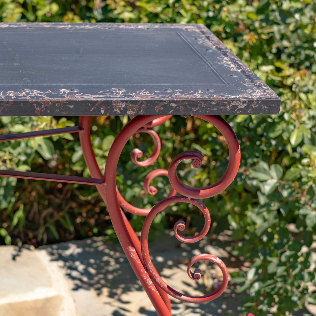 Close view of a dark rectangular tabletop with worn edges above an ornate red iron scrollwork leg.