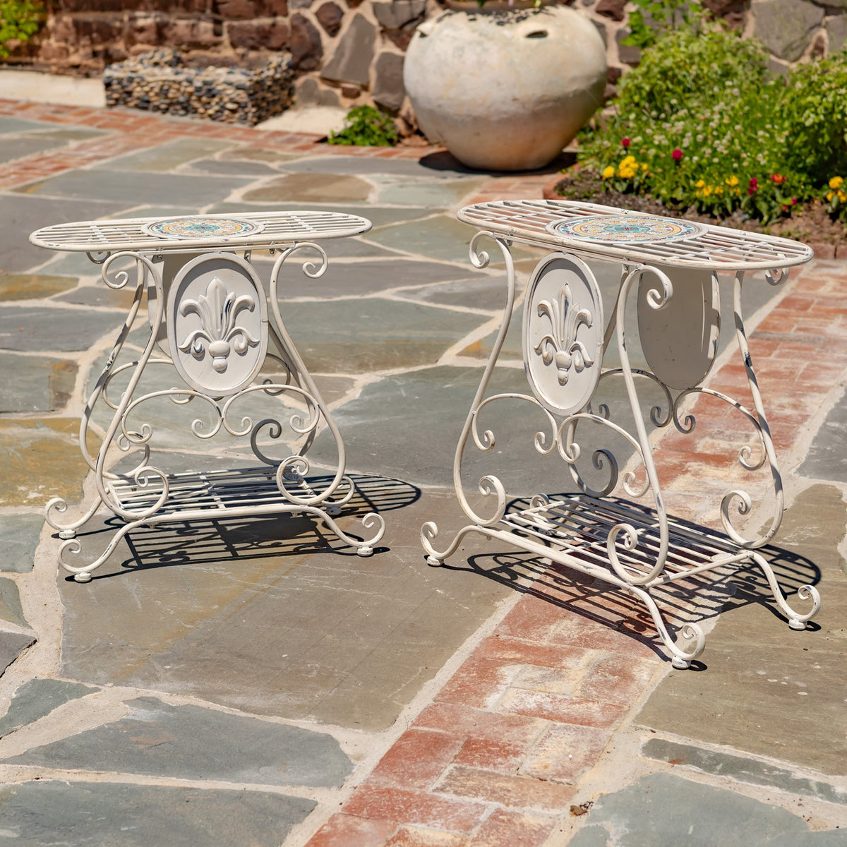 Two matching cream plant stands shown together on a stone patio, with mosaic insets and decorative scrollwork.