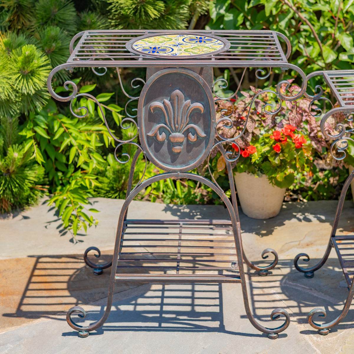 Close view of a plant stand's colorful mosaic tabletop, fleur-de-lis ornament, and curled metal legs.