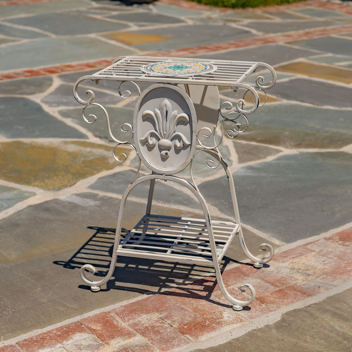 An ornate plant stand viewed from the front, with a round emblem and slatted lower shelf.