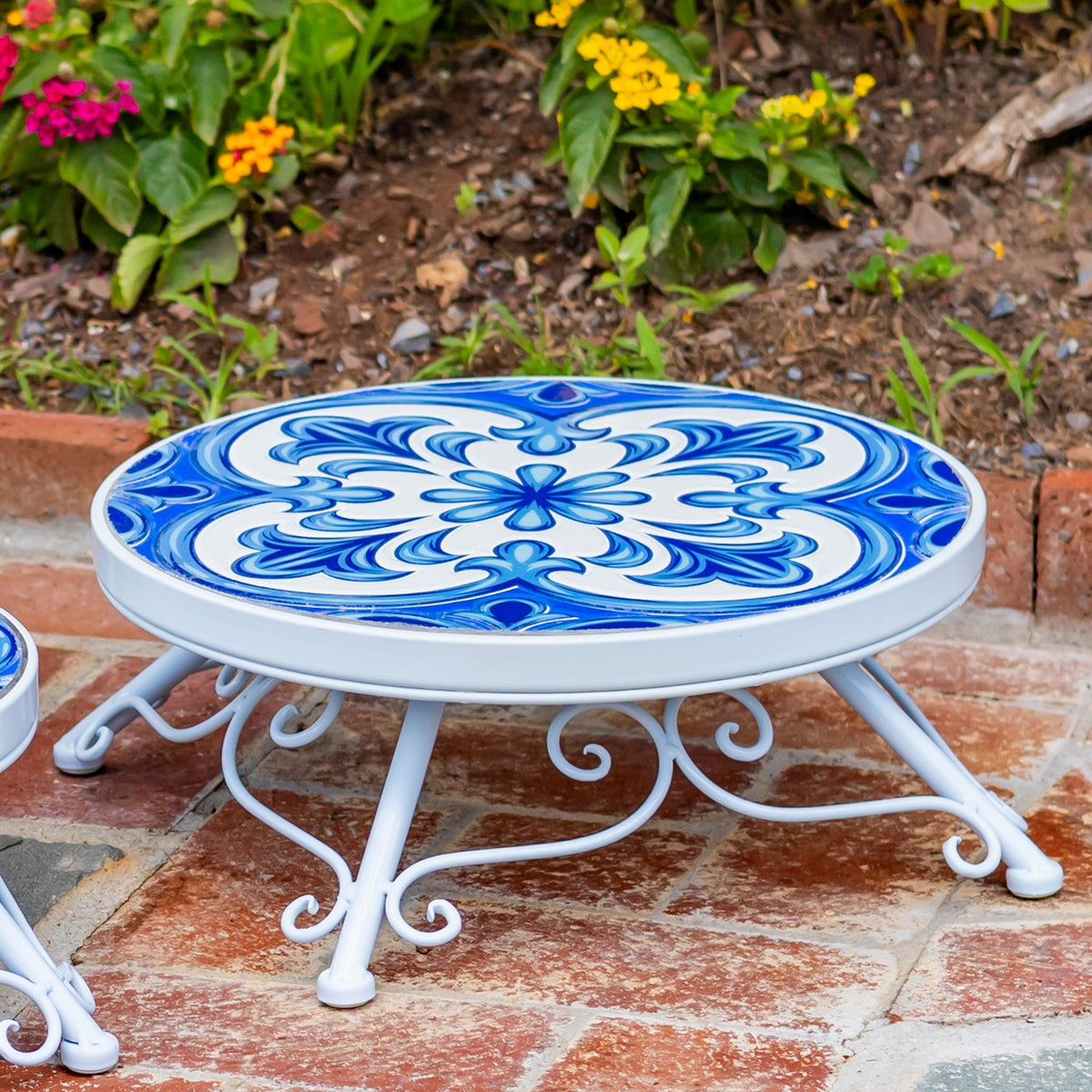Close view of a white metal stand with a blue patterned top, set among patio tiles and garden plants.