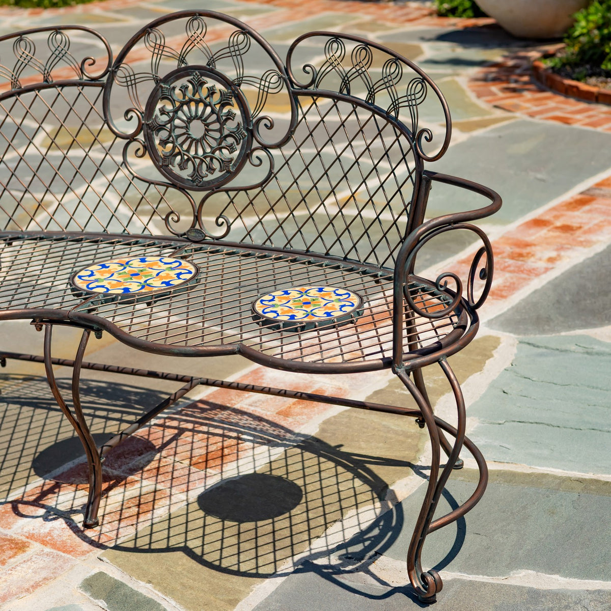 Close view of ornate metal back scrollwork and a curled arm above the mosaic-tiled bench seat.