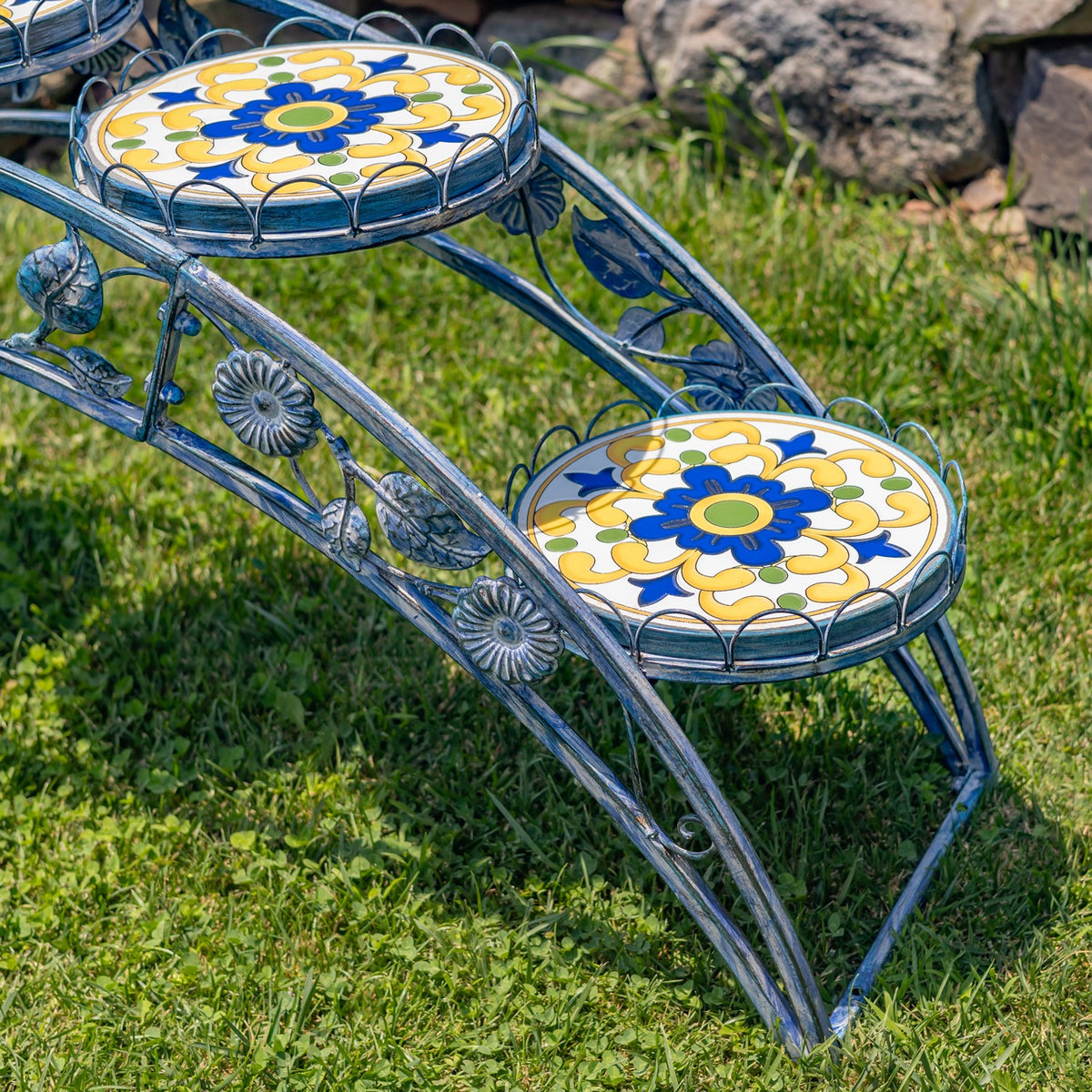 Close-up of two round blue-and-yellow floral mosaic platforms and the stand’s curved iron frame.