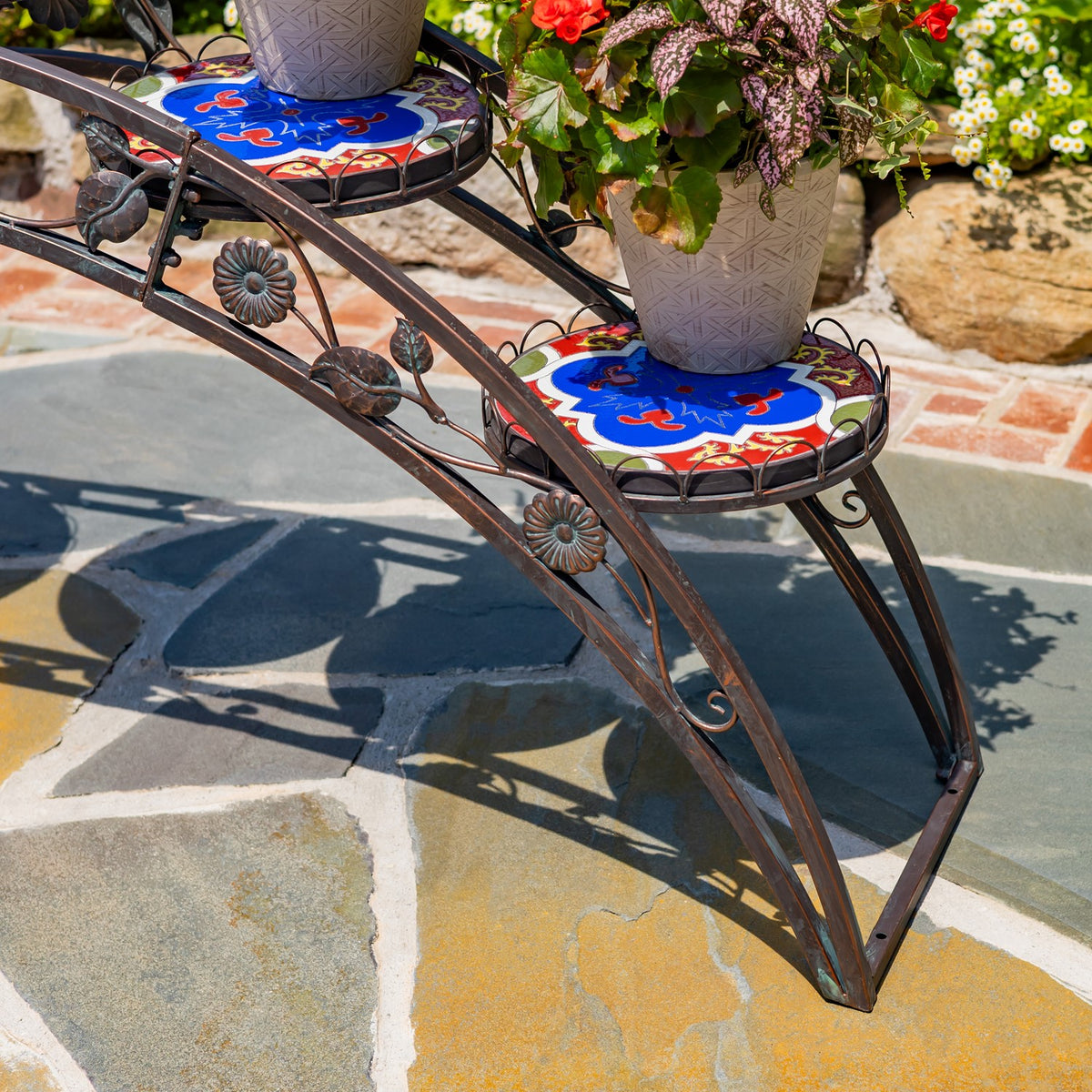 Close-up of patterned mosaic platforms, potted flowers, and floral iron accents on the stand.
