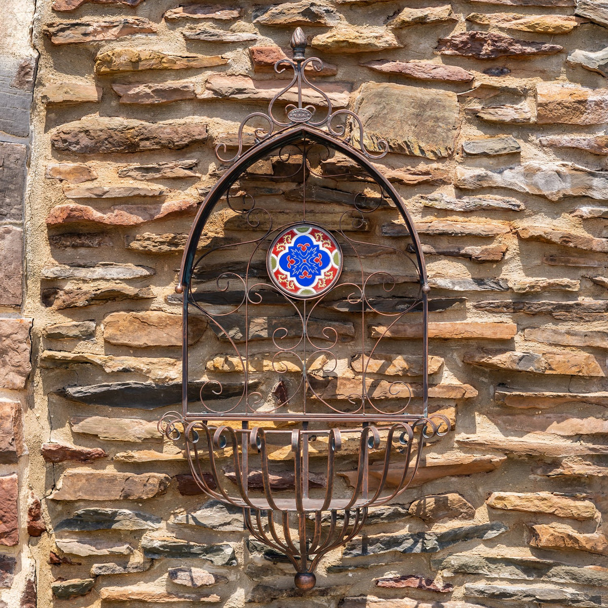 Ornate bronze-toned wall planter with an empty curved basket and a colorful tile medallion against stone.