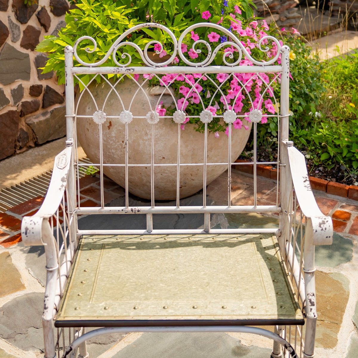 White metal chair with an ornate scrollwork back and distressed pale seat, shown on a stone patio.