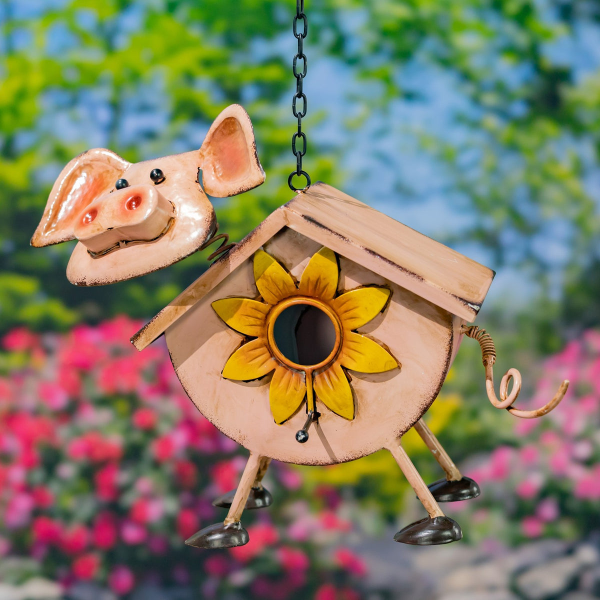 Peach pig-shaped metal birdhouse with a sunflower entrance and curled tail, hanging in a garden.