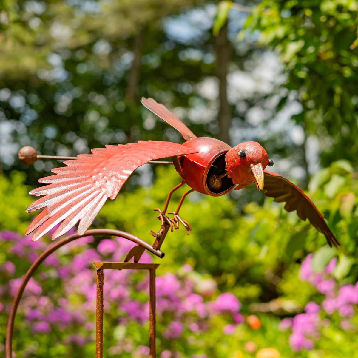 Red metal bird stake with outstretched wings against a flowering garden