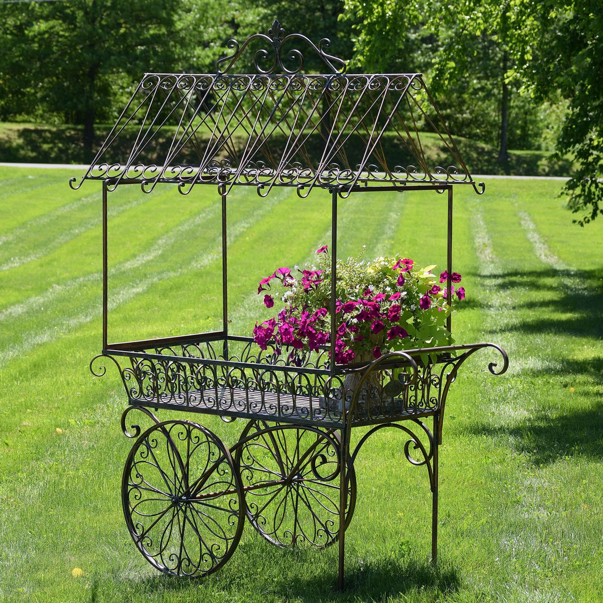Ornate black iron flower cart with pink flowers displayed on a lawn beneath trees.