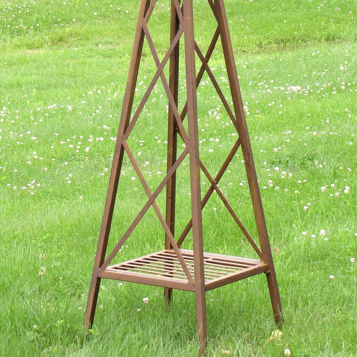 Close view of a rust-brown metal windmill stand with crossed braces and a slatted square base on grass.